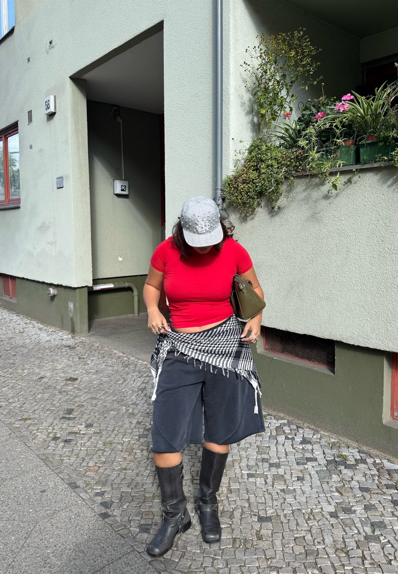 T-shirt rosso attillato, pantaloni larghi neri, cappellino grigio con dettagli, e stivali neri fino al ginocchio. Sciarpa a quadratini bianca e nera legata in vita.