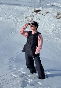 Femme debout dans une neige profonde, portant un pantalon noir, un gilet noir par-dessus une veste polaire rose, une casquette noire et des lunettes de soleil, tenant la visière de la casquette.