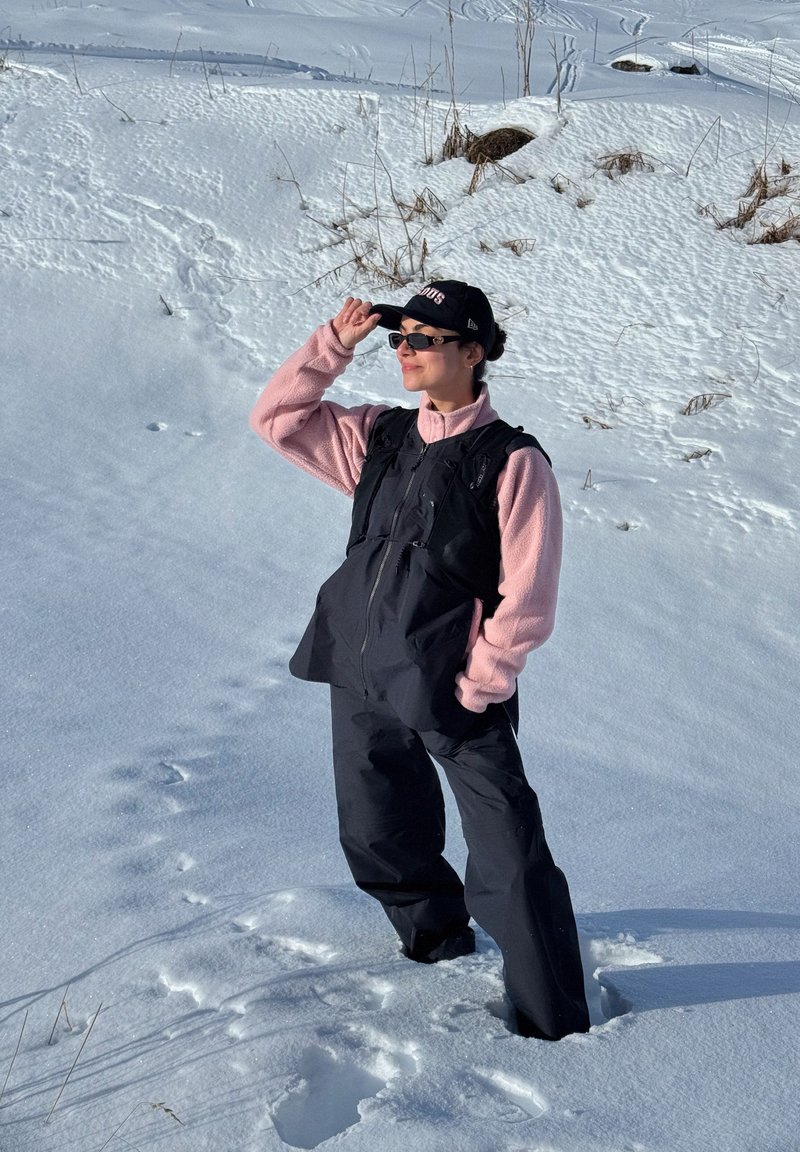 Femme debout dans une neige profonde, portant un pantalon noir, un gilet noir par-dessus une veste polaire rose, une casquette noire et des lunettes de soleil, tenant la visière de la casquette.