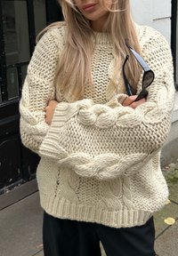 Suéter crema de punto con hilo grueso y texturizado y puños extragrandes, llevado sobre pantalones oscuros, con gafas de sol en la mano.