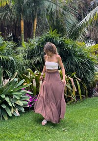 Kobieta w białym topie typu tube i długiej brązowej spódnicy spacerująca po trawie, otoczona tropikalnymi roślinami i fioletowymi kwiatami.