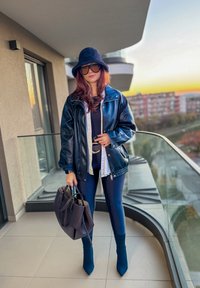 Chaqueta oversize de cuero negro, top negro ajustado y leggings, botines azules de tacón y bolso marrón. Gafas de sol con montura transparente y sombrero de lana.