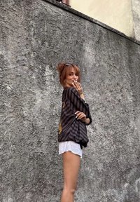 Camisa oversized listrada em preto e dourado, feita de tecido leve, usada com calções brancos. O fundo apresenta uma parede cinza texturizada.