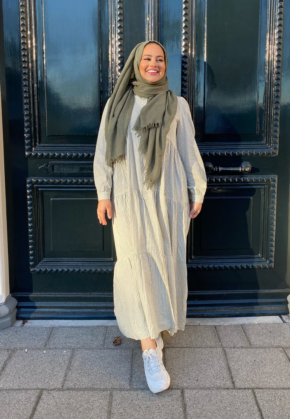 Langes, gestuftes Kleid mit vertikalen weißen und hellgrünen Streifen, kombiniert mit einem grauen Schal und weißen Sneakern, vor einer dunklen Holztür.