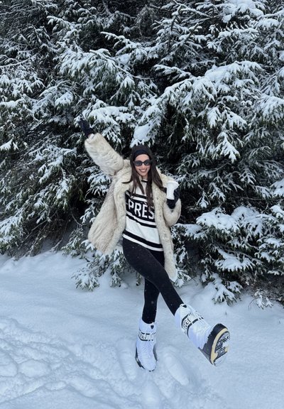 Mujer con abrigo de piel, gafas de sol y botas de nieve sonríe, sosteniendo una taza, de pie sobre la nieve con árboles perennes nevados al fondo.