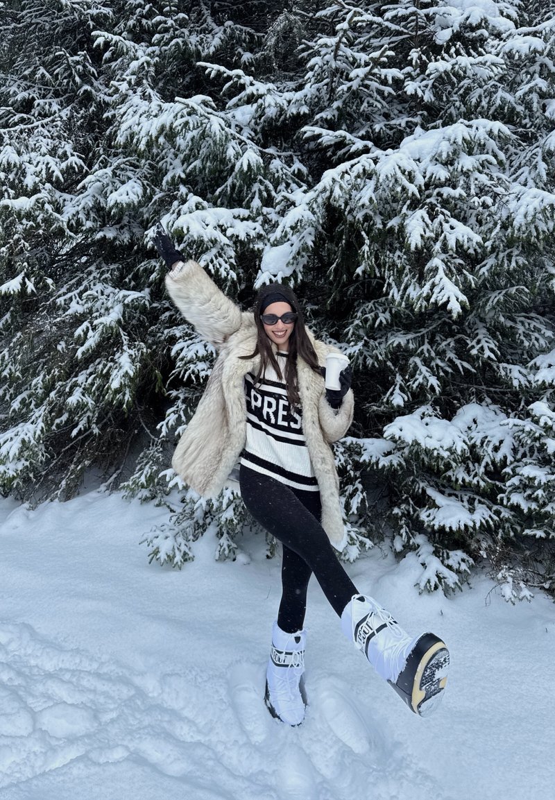 Femme en manteau de fourrure, lunettes de soleil et bottes de neige souriante, tenant une tasse, debout sur la neige avec des arbres à feuilles persistantes enneigés en arrière-plan.