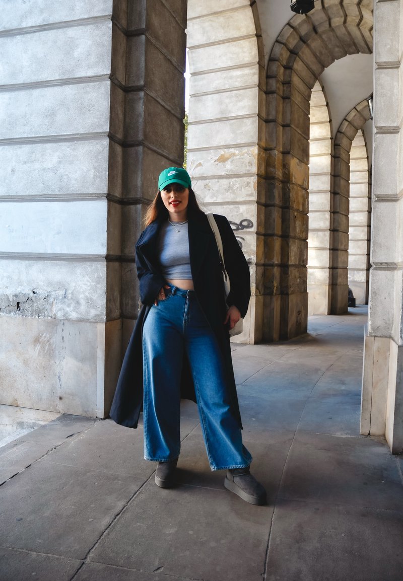 Μαύρο παλτό, γκρίζο κοντό τοπ, μπλε παντελόνια με φαρδιά πόδια, πράσινο καπέλο και γκρι μποτάκια μέχρι τον αστράγαλο; στέκεται σε μια καμάρα με πέτρινους τοίχους.