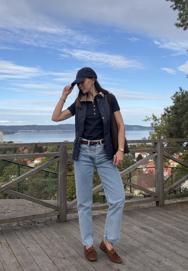 Mujer con ropa informal y gorra de pie en una terraza de madera con vistas al agua y a la ciudad bajo un cielo azul parcialmente nublado.