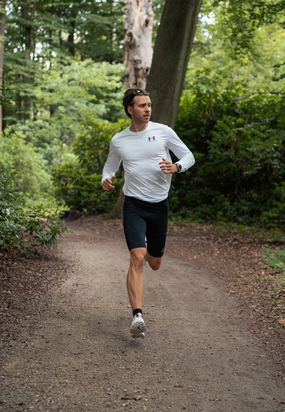 Un atleta con una camiseta de manga larga blanca y pantalones cortos negros corre por un sendero de tierra rodeado de vegetación, llevando un reloj y zapatillas de correr.