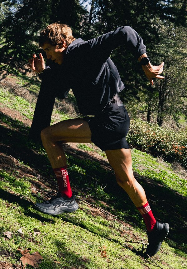 Mustas spordiriietuses jooksja punaste sokkidega jookseb päikeselises metsas ülesmäge sprinti.