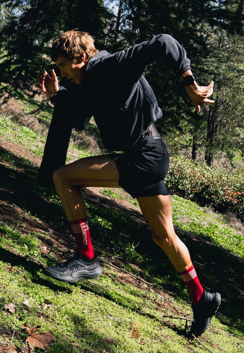 Løper i svart treningstøy og røde sokker spurter oppover en solfylt skogkledd bakke.