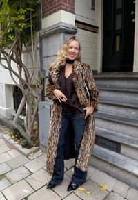 Kvinne med langt blondt hår iført leopardmønstret kåpe, svart topp og jeans, stående på fortauet nær bygningstrapper og et tre.