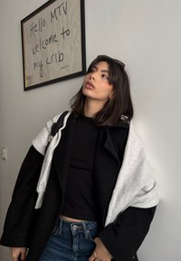 Jeune femme en haut noir, jean et vestes superposées regardant vers le haut, debout à côté d'un panneau encadré sur lequel est écrit « Hello MTV welcome to my crib ».