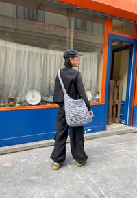 Szürke kötött válltáska, texturált, vaskos dizájnnal. Fekete nadrággal és egy megfelelő fekete kabáttal viselve, lapos sapkával.