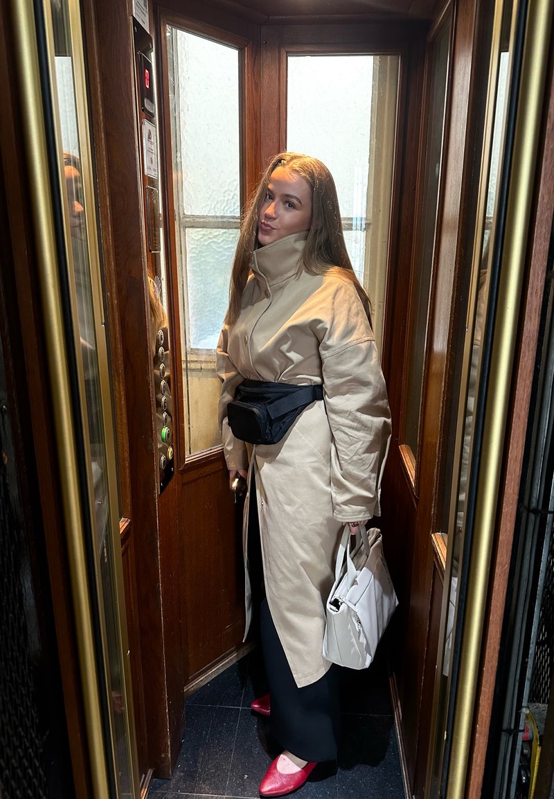 Mujer con abrigo beige, pantalones negros, zapatos rojos y bolso negro de cintura, de pie dentro de un ascensor con paneles de madera sosteniendo un bolso blanco.