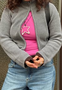 Pull gris à fermeture éclair à côtes avec un col haut, porté par-dessus une chemise rose. Comprend des poignets marron et un collier pendentif en forme d'étoile. Jean bleu clair visible.