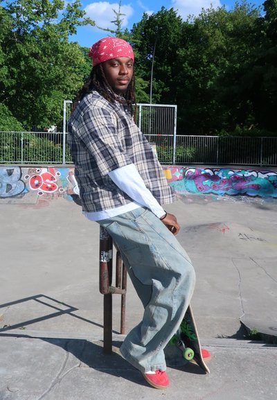 Camisa de cuadros de manga corta, camiseta blanca de manga larga debajo, jeans sueltos de color azul claro, zapatillas rojas y un pañuelo rojo. Patineta al lado.