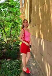 Rosa ribbet tanktopp, røde skreddersydde shorts, røde hæl sandaler med blomsterdetaljer, og en mønstret rød veske; grønne planter i bakgrunnen.