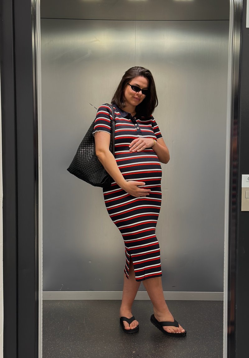 Femme enceinte portant une robe rayée, des lunettes de soleil et des sandales, se tient dans un ascenseur, tenant son ventre avec un grand sac noir à épaule.