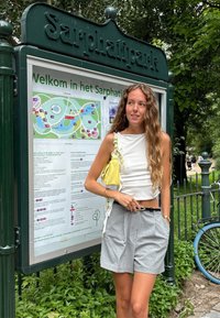 Top bianco senza maniche, pantaloni corti plissettati grigio chiaro, bracciale. Tiene una borsa gialla. Segnaletica del parco verde sullo sfondo.