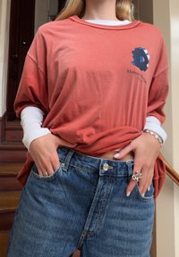 Άτομο που φορά στρωμένα πορτοκαλί και λευκά μακριά μανίκια με μπλε τζιν, στέκεται με τα χέρια στις τσέπες, δείχνοντας ασημένια δαχτυλίδια και βραχιόλι.