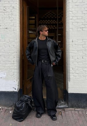 Chaqueta de cuero negra sobre una camiseta negra, combinada con jeans oscuros anchos y zapatillas negras. Una bolsa de deporte oscura descansa en el suelo.