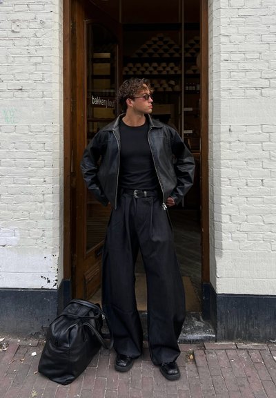 Chaqueta de cuero negra sobre una camiseta negra, combinada con jeans oscuros anchos y zapatillas negras. Una bolsa de deporte oscura descansa en el suelo.