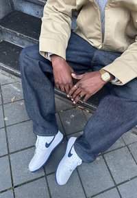Witte Nike Air Force 1 sneakers met marineblauwe accenten, gecombineerd met donkerblauwe losse jeans en een beige ritsvest. Gouden horloge om de pols.