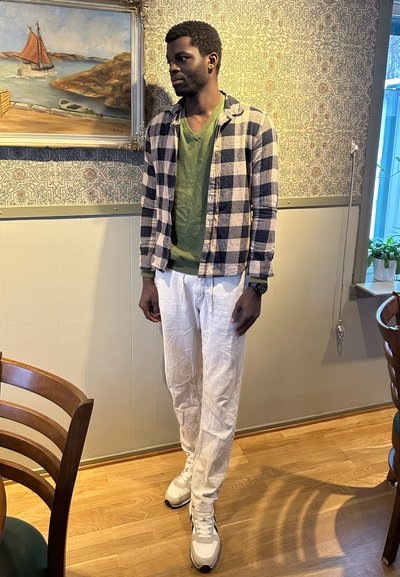 Hombre con camisa verde, chaqueta a cuadros y pantalones blancos, de pie en el interior junto a una pintura enmarcada de un barco junto al agua.