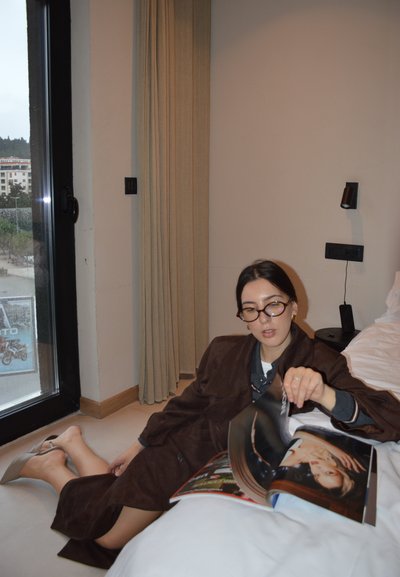 Mujer con gafas y abrigo marrón sentada en el suelo junto a la cama, hojeando una revista de moda en una habitación de hotel cerca de una puerta de cristal.
