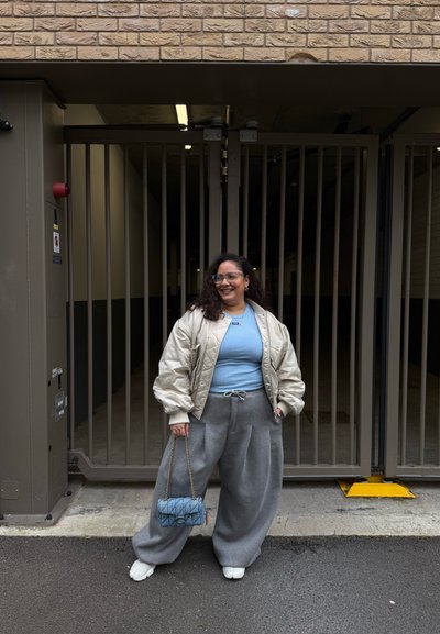 Mujer sonriente con gafas frente a una entrada con reja, lleva una chaqueta ligera, top azul, pantalones anchos grises, zapatos blancos y sostiene un bolso azul.