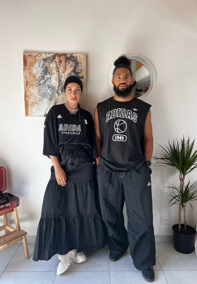 Dos personas llevan ropa negra de Adidas. Una lleva una falda larga negra y una camiseta, mientras que la otra lleva una camiseta sin mangas negra y pantalones holgados. Se ven zapatillas blancas y negras.