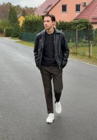 Zwarte leren jas, donkere sweater, lichtgrijze broek en witte sneakers; wandelend op een weg met groen en huizen op de achtergrond.