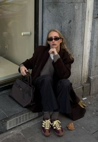 Femme aux longs cheveux blonds portant des lunettes de soleil, un manteau sombre, un pantalon à fines rayures et des baskets, assise sur un trottoir tenant un grand sac à main foncé.