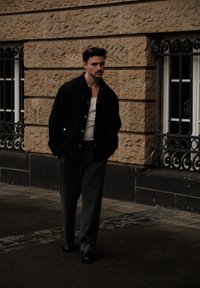 Svart jakke med knapper over en hvit skjorte, grå bukser og svarte sko. Bakgrunnen viser en steinvegg og et vindu med utsmykket jernarbeid.