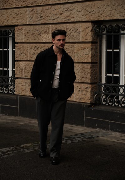Chaqueta negra abotonada sobre camisa blanca, pantalones grises y zapatos negros. El fondo muestra una pared de piedra y una ventana con rejas ornamentales de hierro.