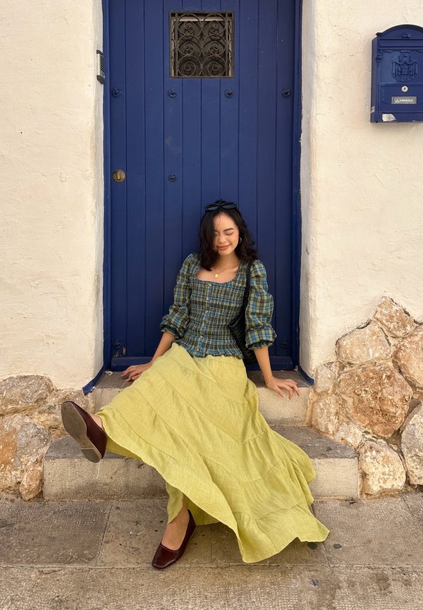 Femme aux cheveux noirs assise sur des marches en pierre devant une porte bleue, portant un chemisier à carreaux verts, une jupe jaune et des chaussures marron.