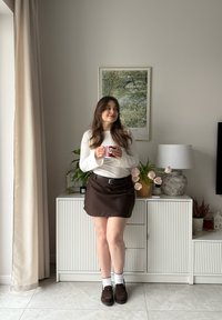 Hvit langermet topp, brun stripet skjørt med belte, kombinert med hvite ankelsokker og brune loafers. Innendørs setting med planter.