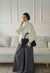 Femme en pull blanc et pantalon large foncé tenant un sac à main noir, debout à l'intérieur près d'un canapé beige contre un mur blanc.