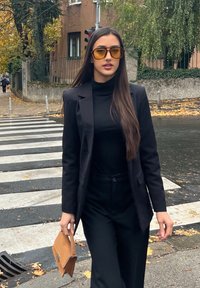 Must musta bleiser üle kõrge kaelusega pluusi, kombineeritud mustade pükstega. Suured kollased päikeseprillid ja pruun käekott. Ülekäigurada ja sügislehed on näha.