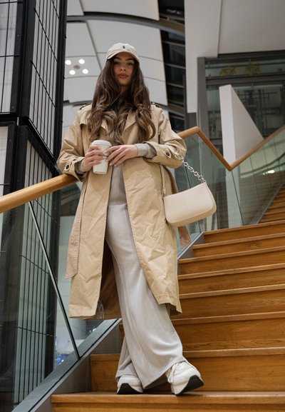 Trench beige, top gris y pantalones anchos blancos. Sosteniendo una taza de café, con una gorra beige. Pequeño bolso beige con detalle de cadena. Escaleras de madera.