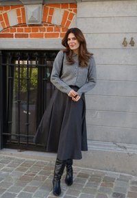 Grauer Strickcardigan mit Knöpfen, kombiniert mit einem langen, dunkelgrauen Rock. Schwarze kniehohe Stiefel vervollständigen das Outfit. Im Hintergrund sind Ziegel und Stein zu sehen.