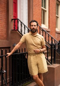 Beige pikétröja och matchande shorts, båda med en slät textur. Modellen lutar sig mot svarta järnräcken med en röd tegelbakgrund.