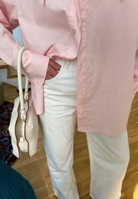 Camisa rosa clara de botão com mangas longas, calças de denim brancas e uma mala creme com zíper dourado e acabamento castanho.