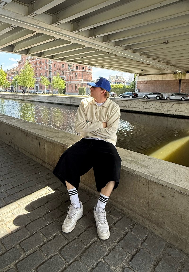 Person iført en beige langærmet skjorte og sorte løse shorts, siddende på en betonafsat ved vandet, med hvide sneakers og stribede sokker.