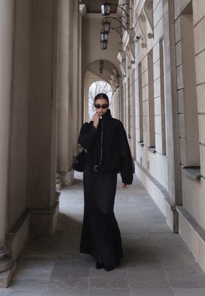 Mujer con falda larga negra y chaqueta con gafas de sol caminando por un pasillo de piedra con columnas y faroles colgantes.