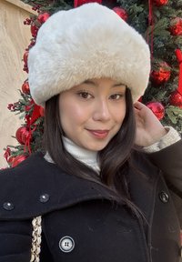 Cappello bianco in pelliccia sintetica sopra un cappotto scuro con bottoni e un dolcevita a coste bianco. Sullo sfondo ornamenti rossi e vegetazione verde.