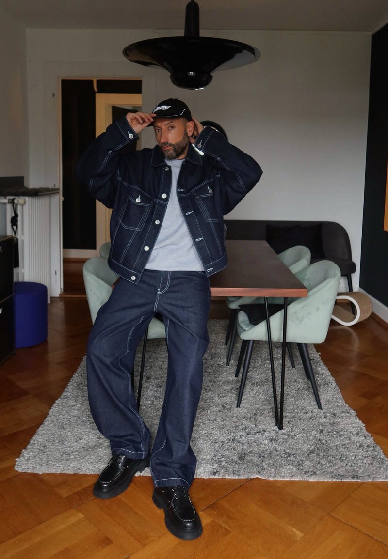 Uomo che aggiusta un cappello nero mentre si appoggia a un tavolo in una moderna sala da pranzo, indossando una giacca in denim scuro e pantaloni con scarpe nere.