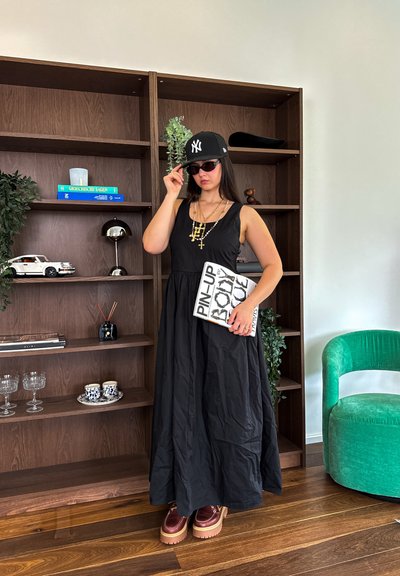 Un vestido maxi negro con un corsé ajustado y una falda fluida, combinado con una gorra negra, gafas de sol y zapatos de plataforma marrones y gruesos. Sostiene un libro.