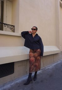 Una camisa cortada de rayas oscuras, una falda midi semitranslúcida con estampado floral en tonos cálidos, calcetines grises y tacones negros puntiagudos.